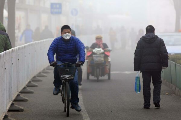 A man wearing a face mask cycling in a smoggy looking street. Another man walking to his right in the other direction and a person on a scooter wearing a face mask in the distance.