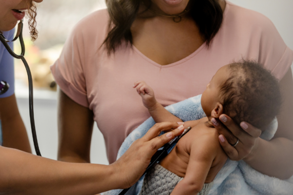 Mother wearing pink shirt holds baby during a medical exam with a stethoscope.